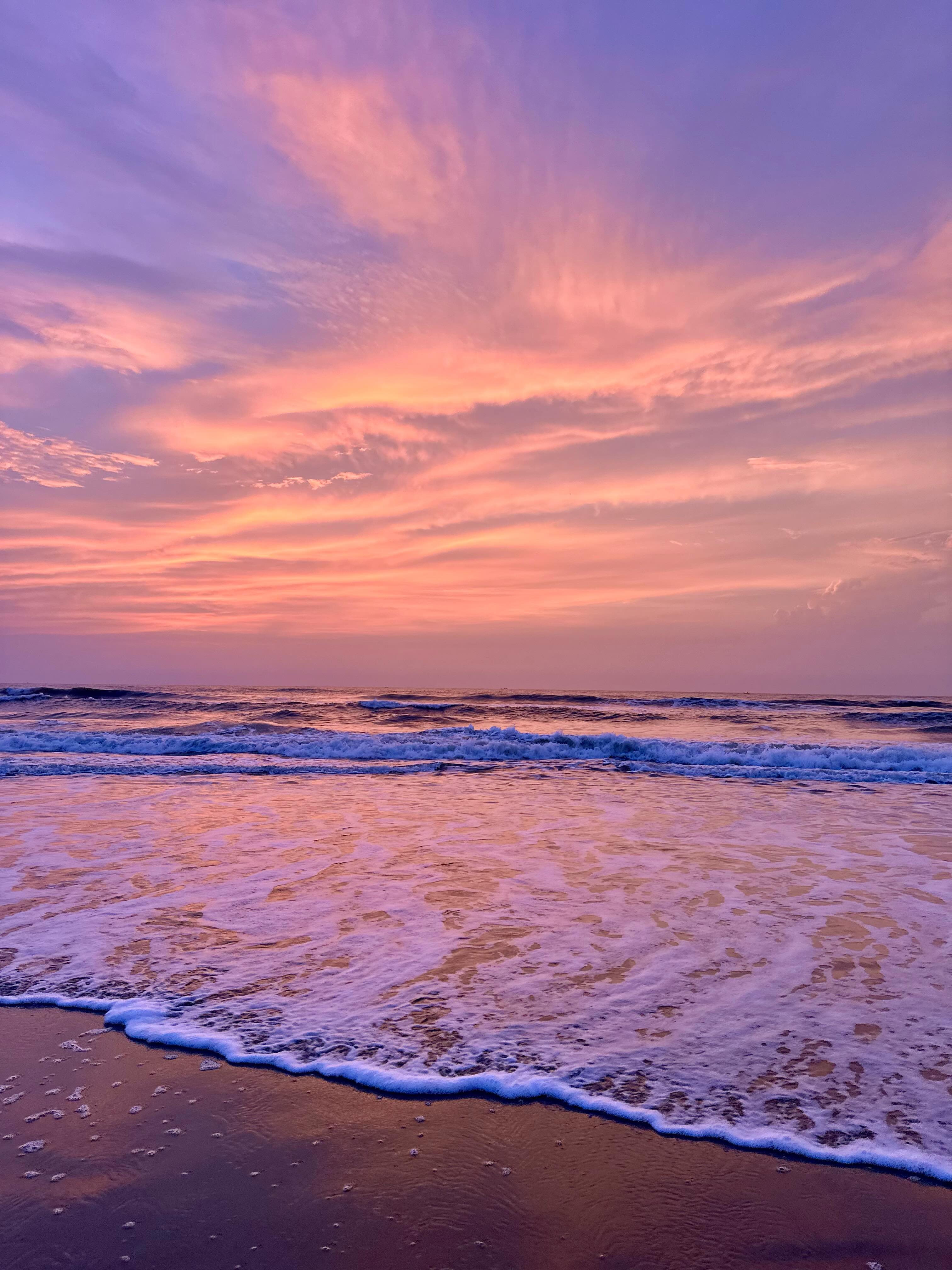 Pink and violet sunset clouds above waves rolling onto a beach.
